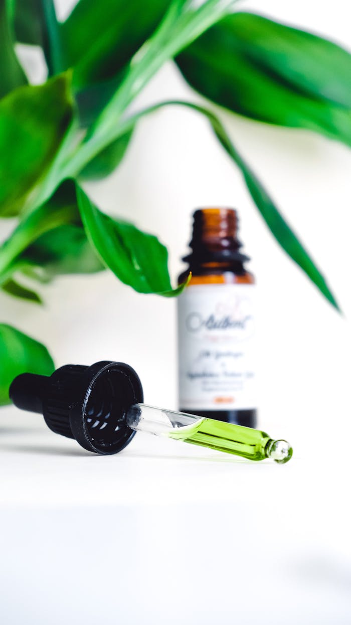 A dropper with green liquid beside a bottle, surrounded by green leaves.